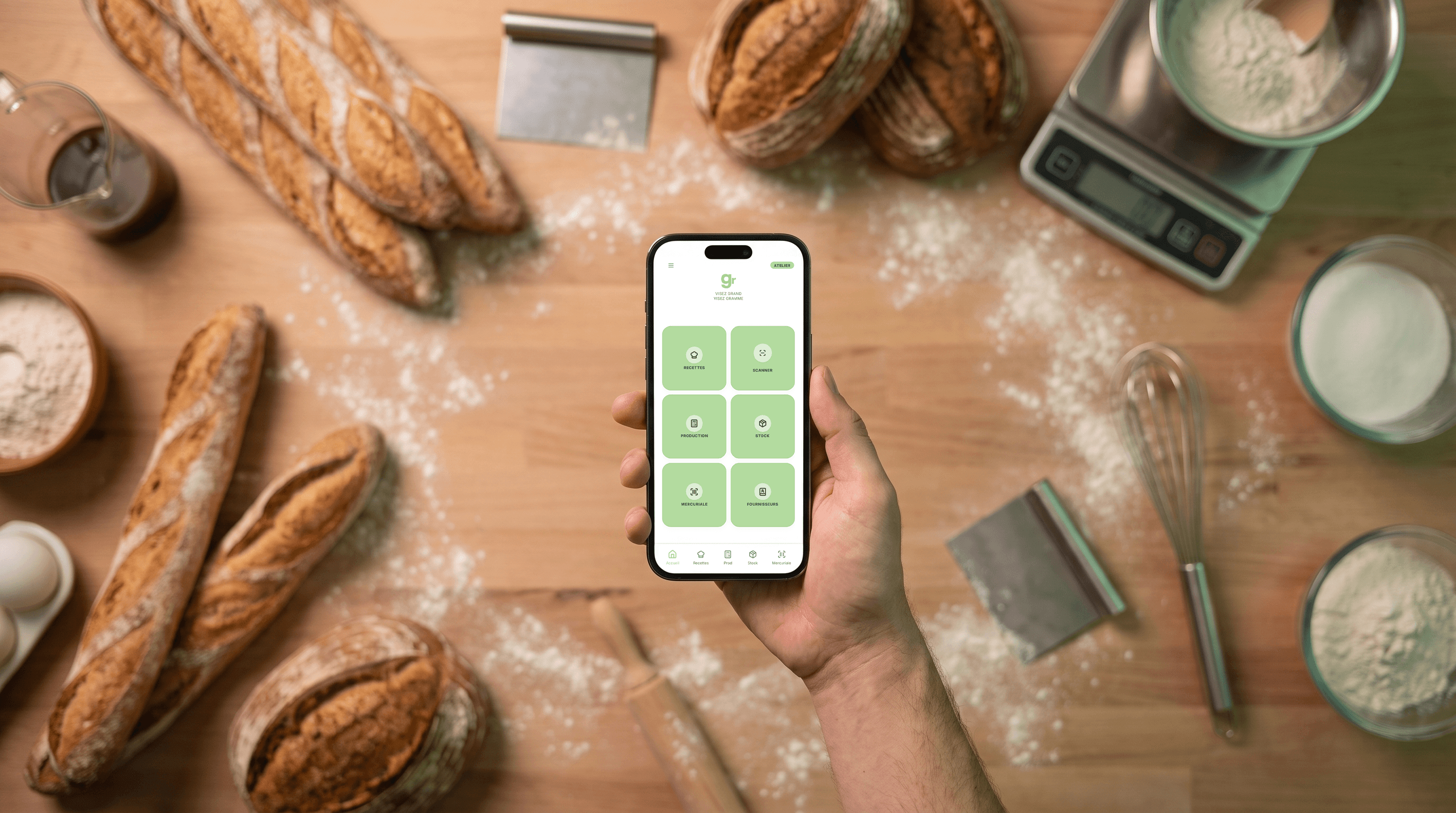 Le logiciel Gramme ouvert sur un téléphone, tenu au-dessus d'un plan de travail avec des baguettes, de la farine et une balance